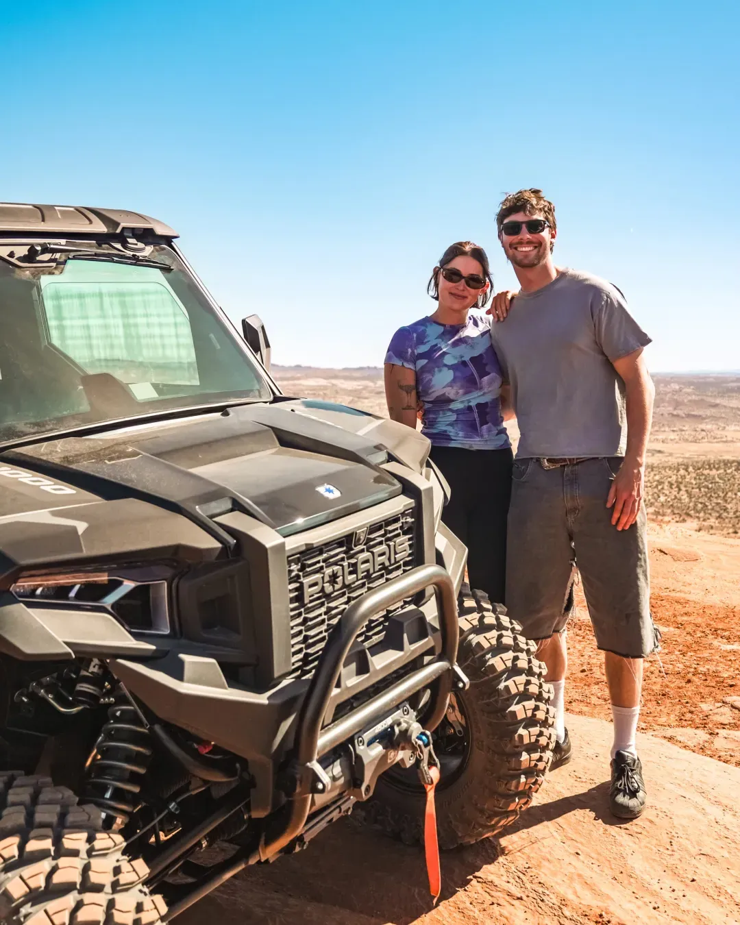 Smiling couple wearing sunglasses standing next to a Polaris off-road vehicle on a rocky terrain with a clear blue sky.