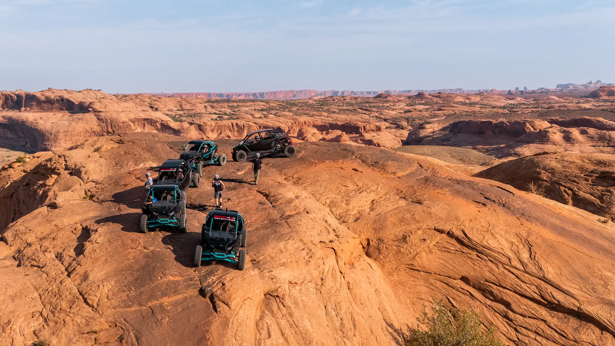 Four off-road vehicles parked on rocky desert terrain with people standing nearby under a clear sky.