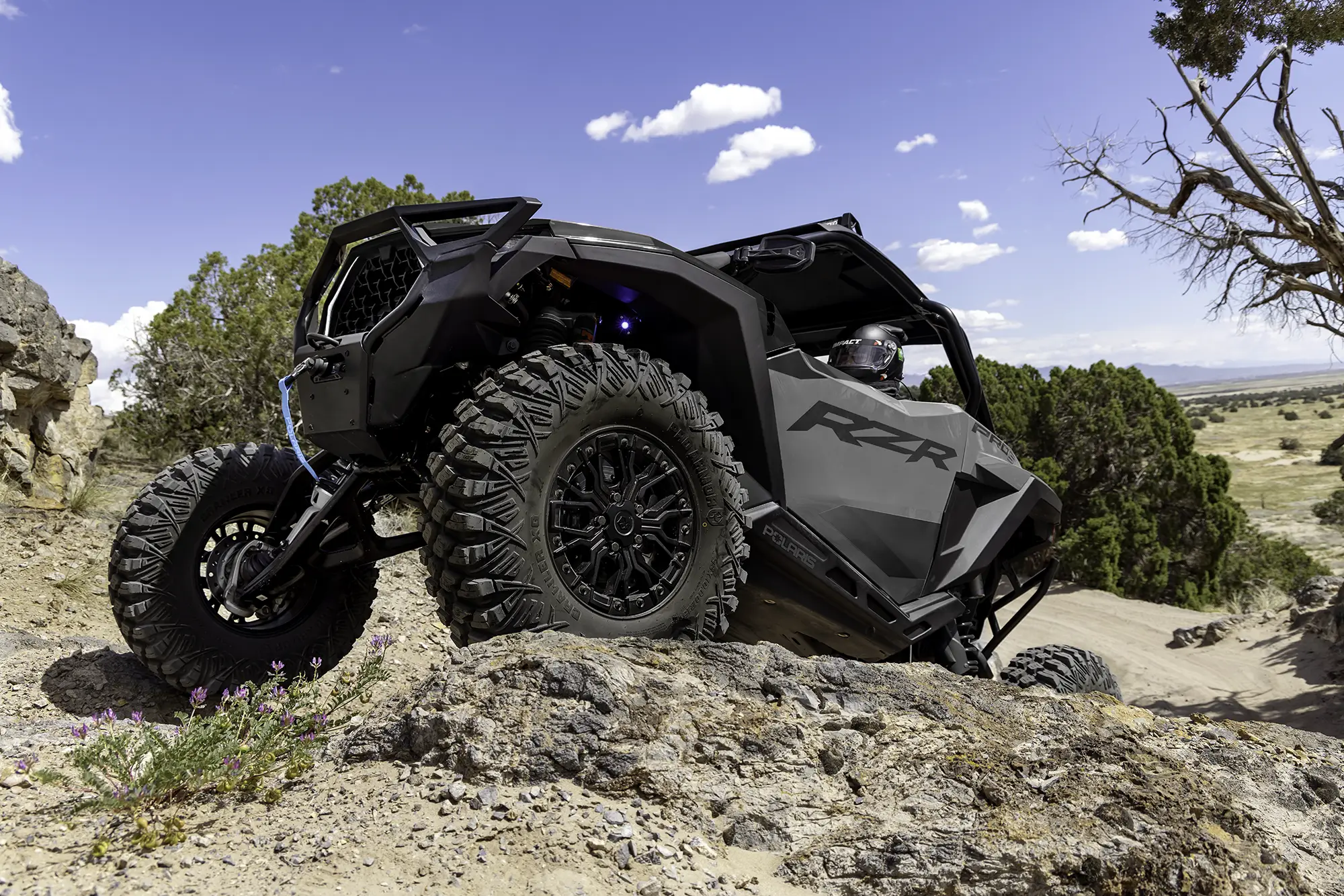 Grey Polaris RZR off-road vehicle climbing rocky terrain with rider wearing a helmet under a blue sky with clouds.