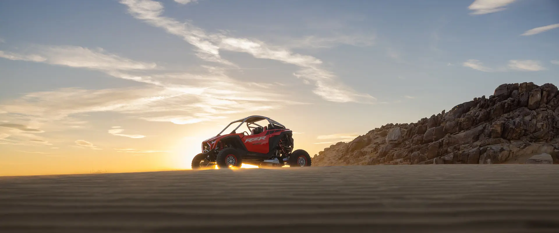 Red off-road vehicle parked on sand dunes with rocky hills in the background at sunset.