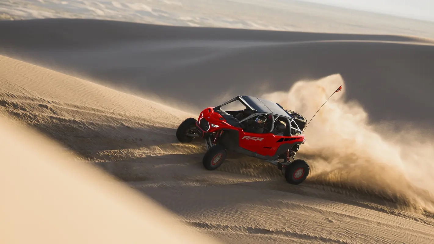 Red off-road side-by-side vehicle kicking up sand while driving over desert dunes.