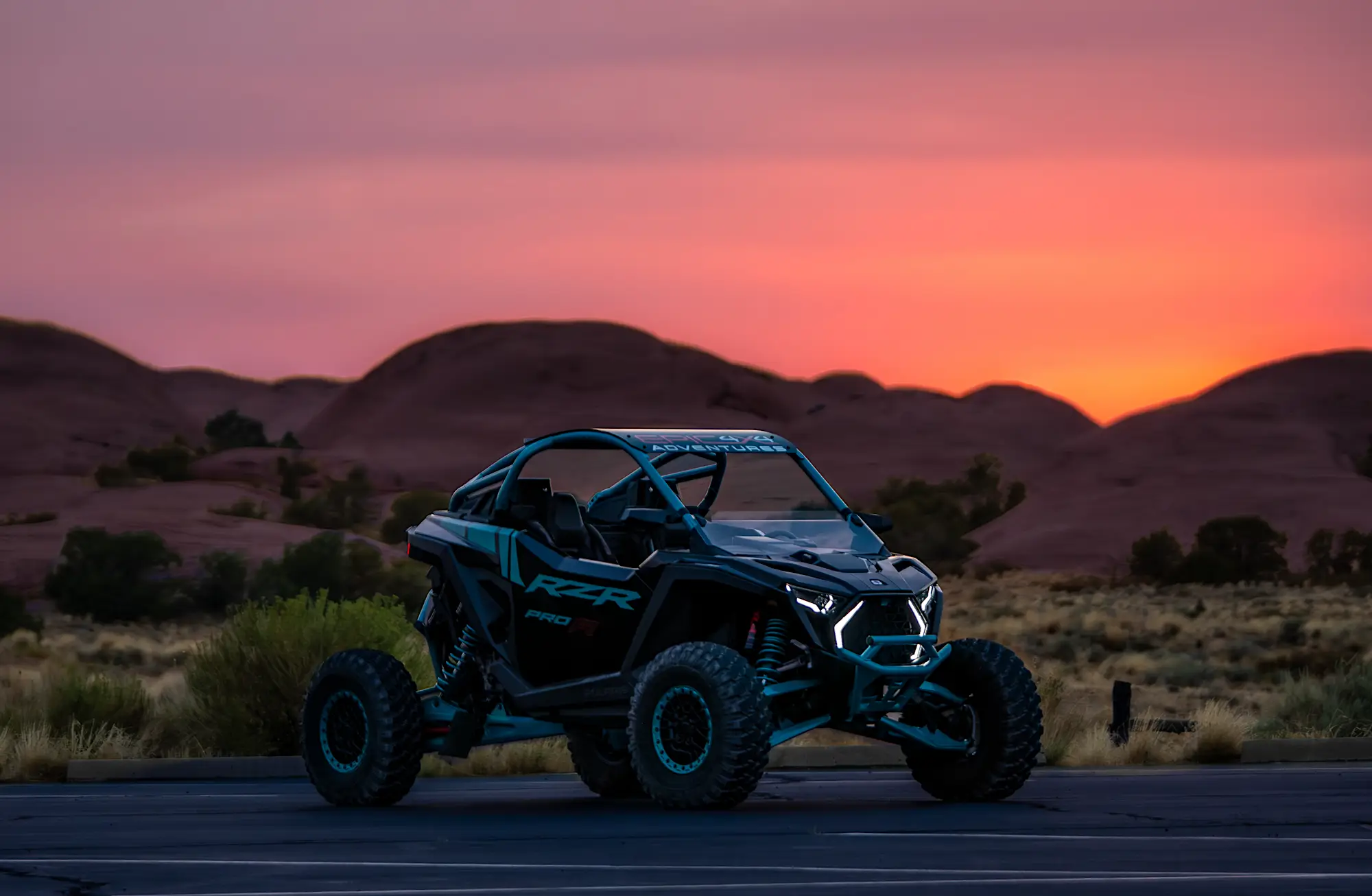 Off-road RZR Pro XP vehicle parked on asphalt with desert hills and an orange-pink sunset in the background.