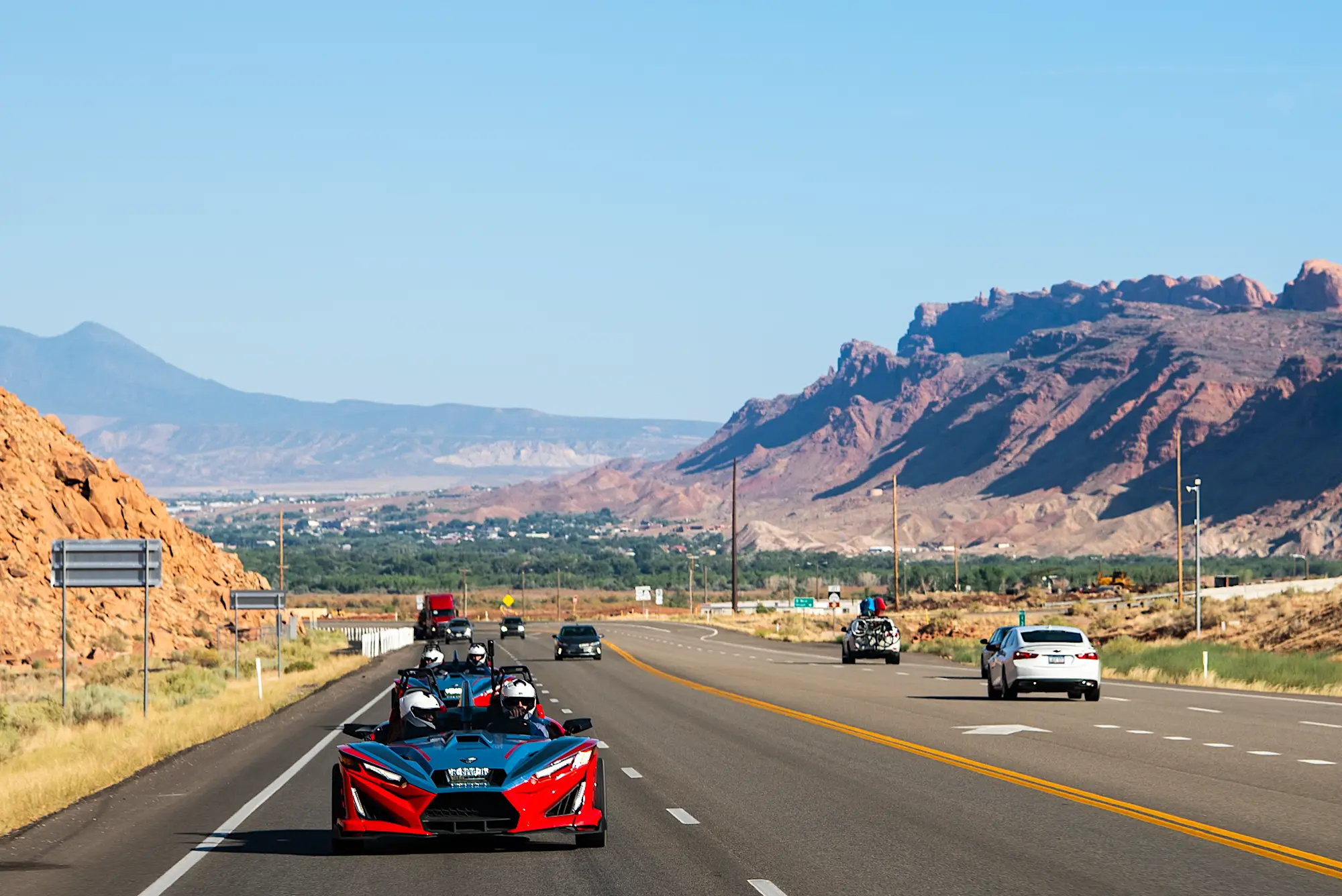Two red and black Polaris Slingshot R driving on a highway with mountainous desert landscape in the background.