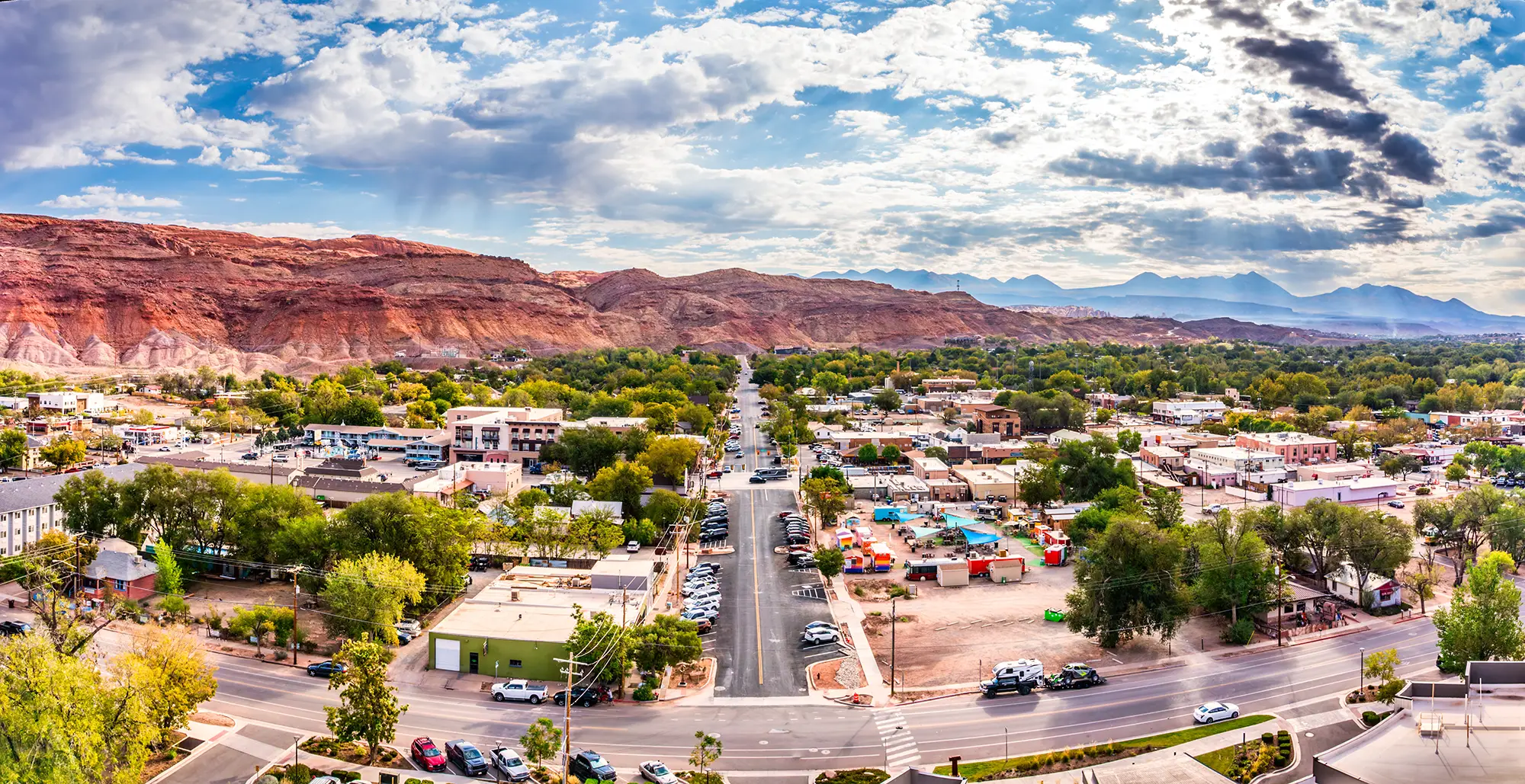 Aerial view of a small town with tree-lined streets and red rock mountains under a partly cloudy sky.