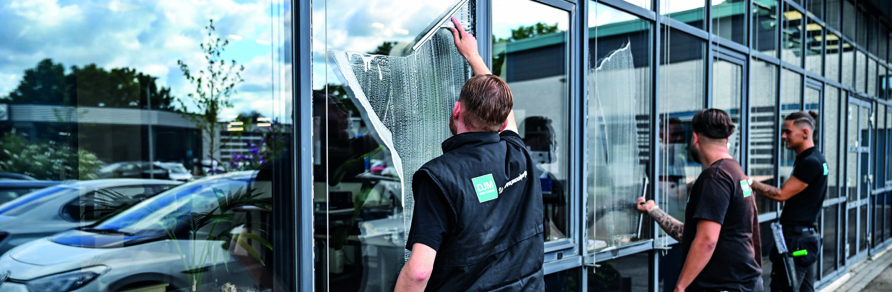 Schoonmaker met een zwarte shirt waarop "DJM – Uw schoonmaakpartner" staat, maakt een glazen deur schoon met een raamwisser, met zichtbare zeepstrepen op het glas.