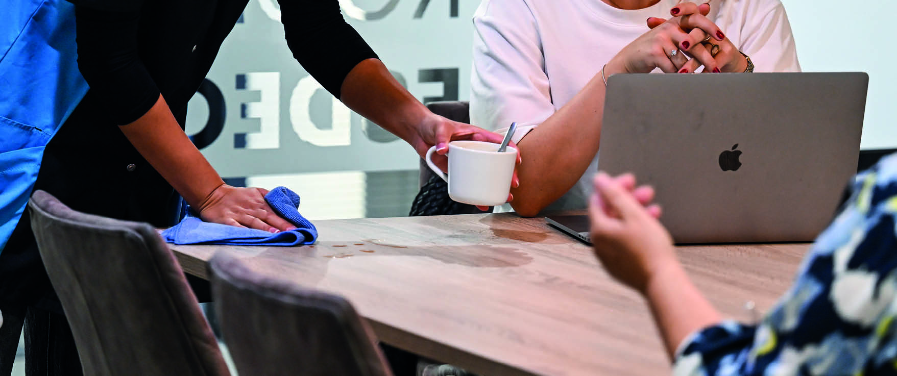 DJM-medewerker maakt een vergadertafel schoon terwijl een kantoormedewerker met laptop en koffiekopje aanwezig is.