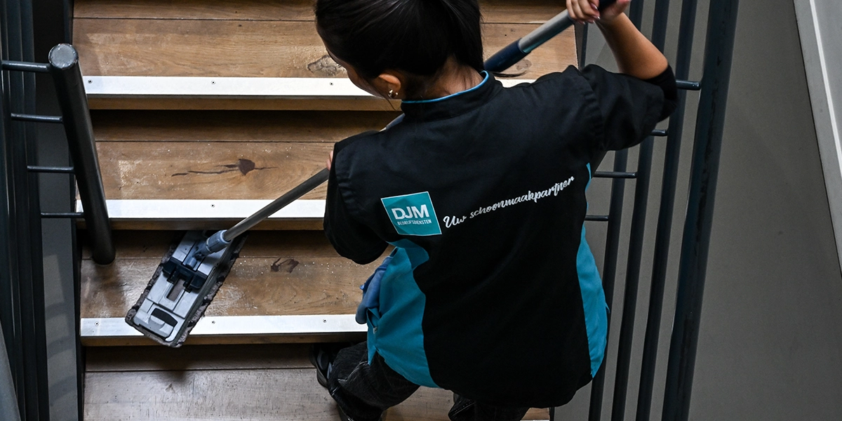 Schoonmaakmedewerker van DJM maakt een houten trap schoon met een vloerwisser, gekleed in bedrijfskleding met het logo en de slogan ‘Uw schoonmaakpartner’.