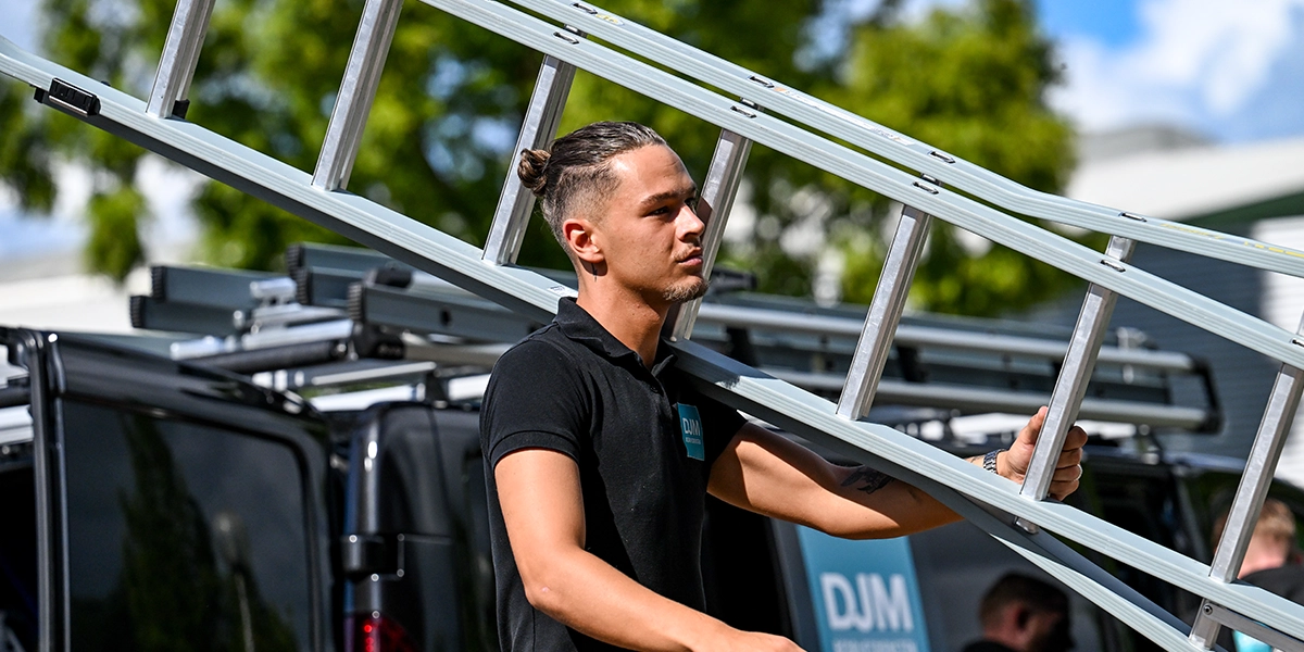 DJM-medewerker draagt een ladder bij een bedrijfsbus met DJM-logo, klaar voor werkzaamheden op locatie in de vastgoedsector.