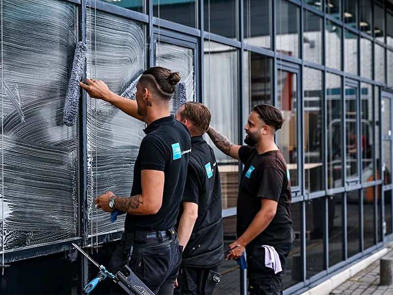 Drie glazenwassers van DJM Gevelreiniging maken samen grote ramen van een bedrijfspand schoon. Ze dragen zwarte werkkleding met het logo van het bedrijf en werken geconcentreerd aan de glanzende gevel.