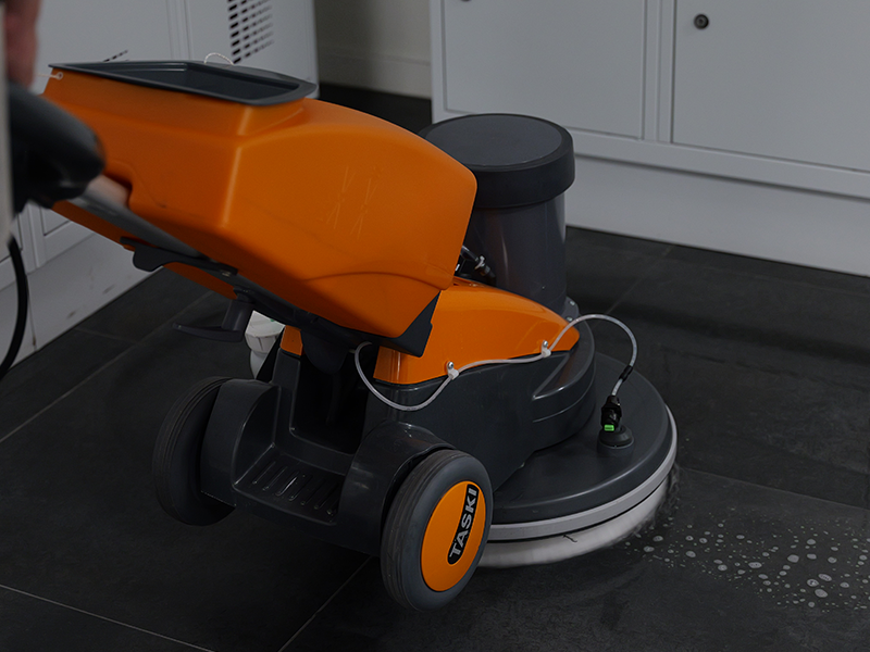 Een oranje schrobmachine maakt de zwarte tegels van een kleedkamer schoon.