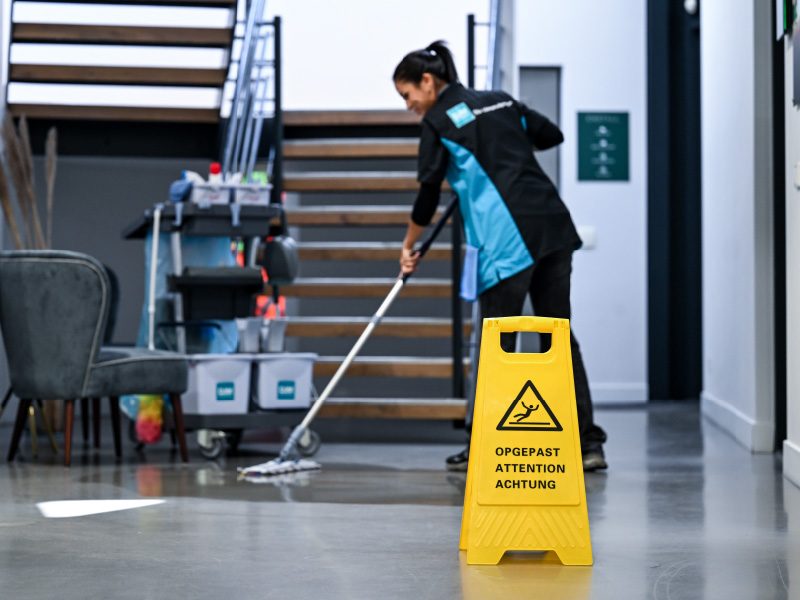 Schoonmaak medewerker dweilt de vloer in een moderne kantoorruimte, met op de voorgrond een gele waarschuwingsbord “Pas op, natte vloer” en op de achtergrond een trap en schoonmaakkar.