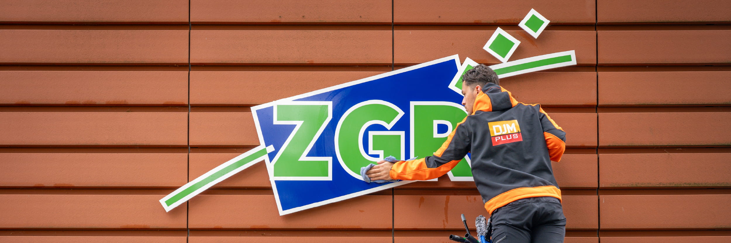 Schoonmaakmedewerker van DJM reinigt een glazen deur met een doek, gekleed in bedrijfskleding met het logo en de tekst ‘Uw schoonmaakpartner’.
