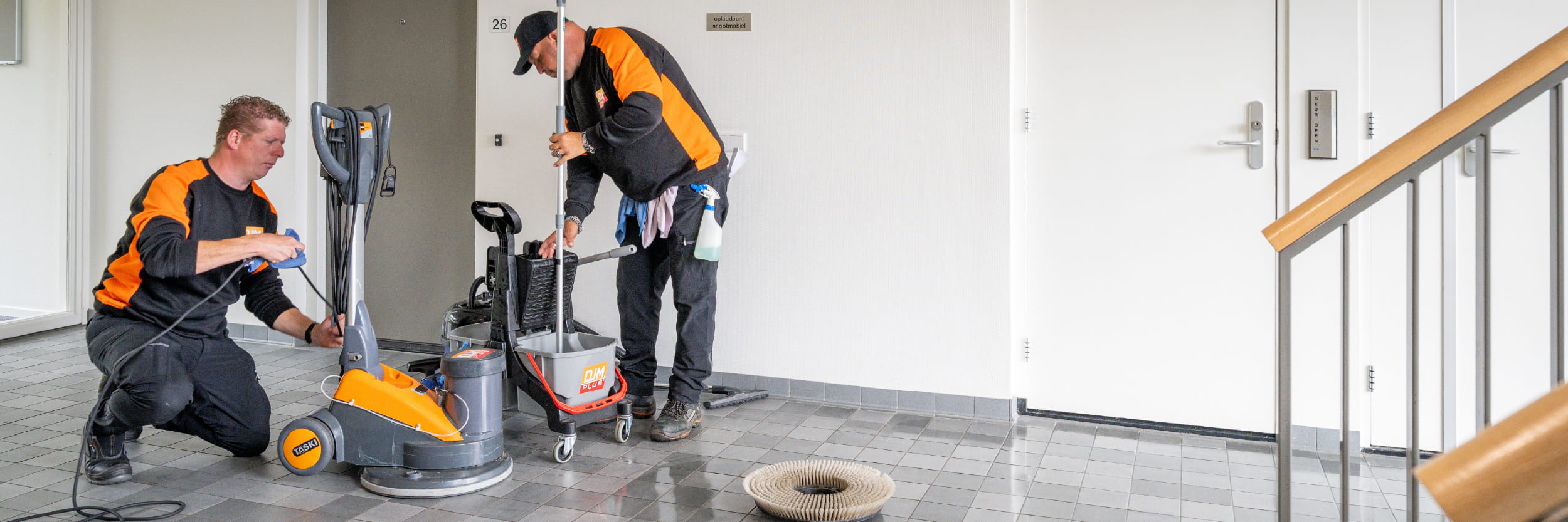 DJM-medewerker maakt de vloer schoon in een industriële opslagruimte, met een schoonmaakkar en bedrijfskleding met DJM-logo.