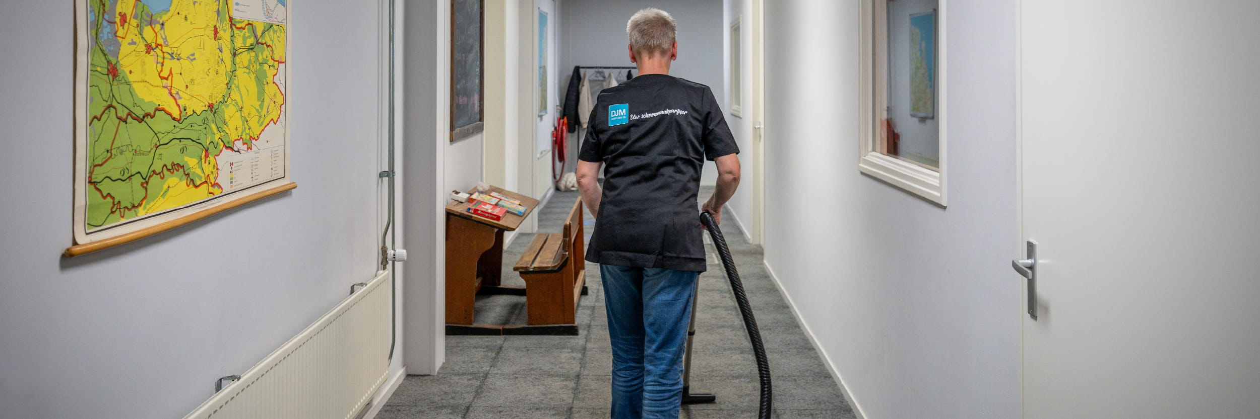 DJM-medewerker stofzuigt horizontale jaloezieën met een kleurrijke plumeau in een onderwijs- of kantooromgeving, gekleed in herkenbare bedrijfskleding.