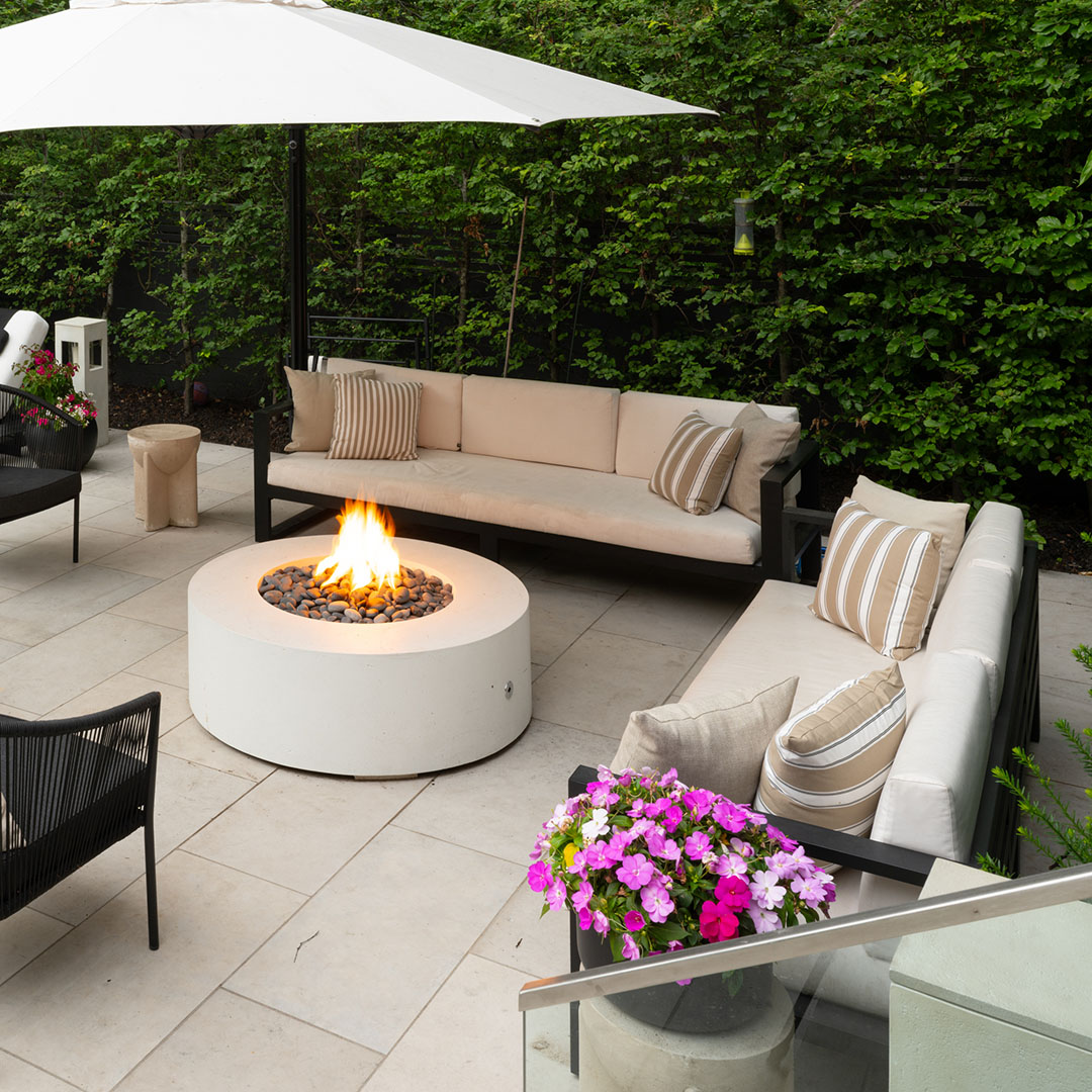 Outdoor patio with fire pit, four chairs with fur throws, and greenery illuminated in the background.