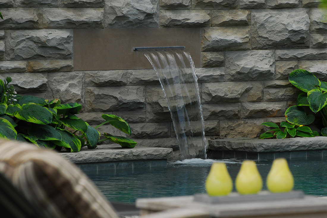 Outdoor seating area with wicker furniture, wooden table, decorative plants, and water feature wall.