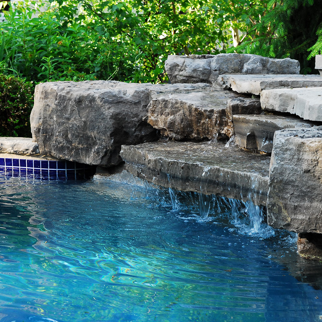 Blue pool next to white stone patio with bench, two lanterns, and green plants surrounding.