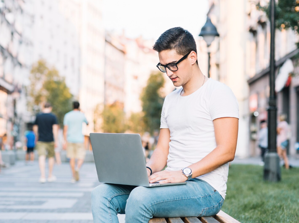 A man wearing glasses and a white shirt, sitting on a bench outdoors and using a laptop