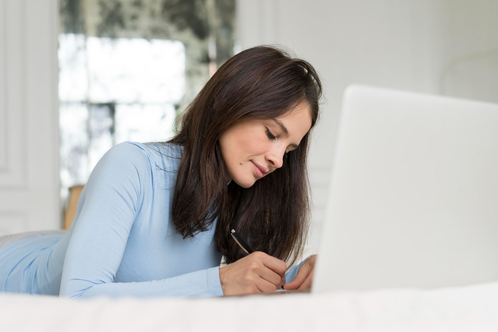 A woman comfortably lying on her bed, researching and writing on her laptop, exploring resources from an addiction treatment website
