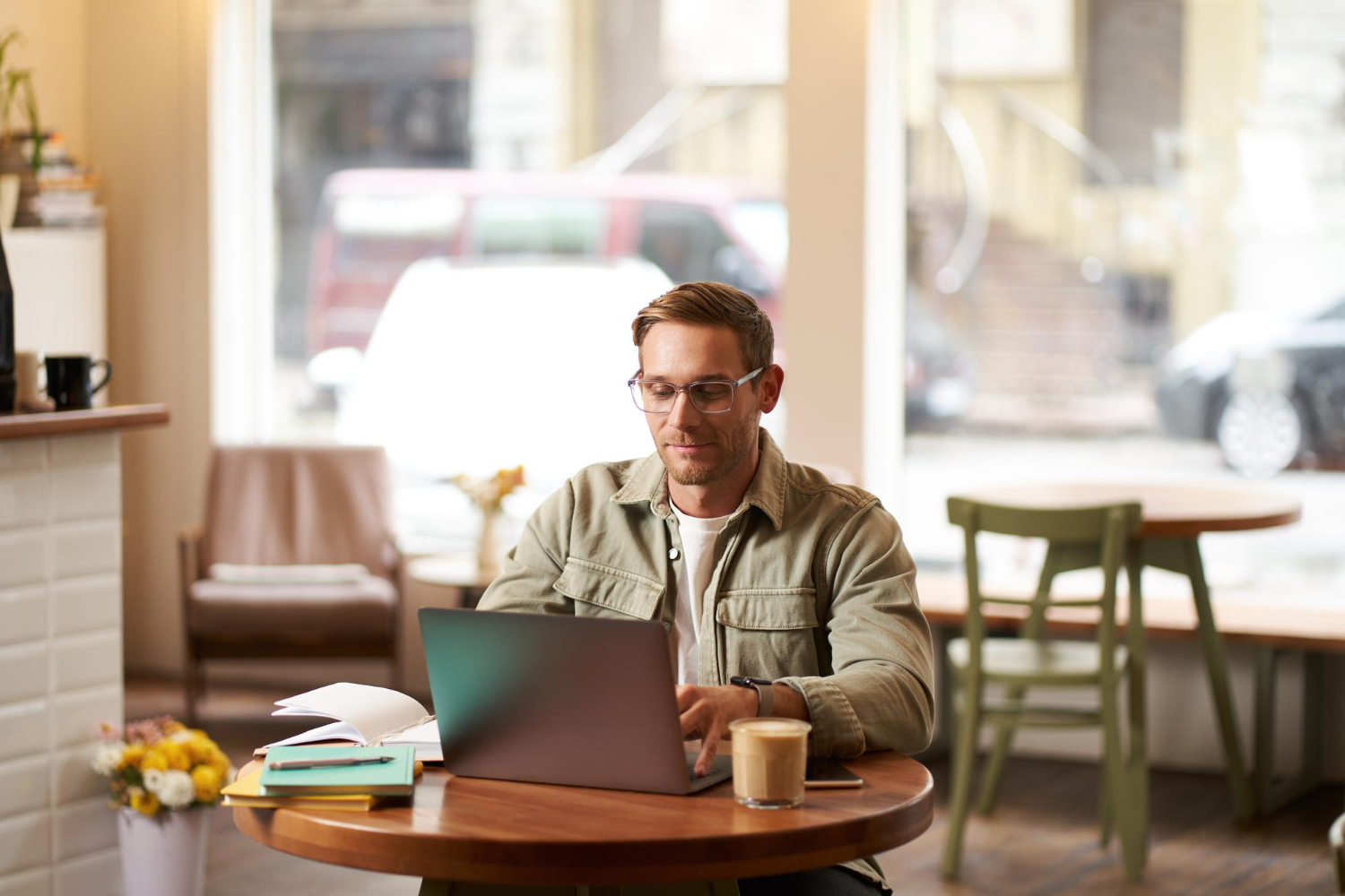 Man wearing glasses working on a laptop at a small café table with notebooks and a cup of coffee, sunlight coming through large windows