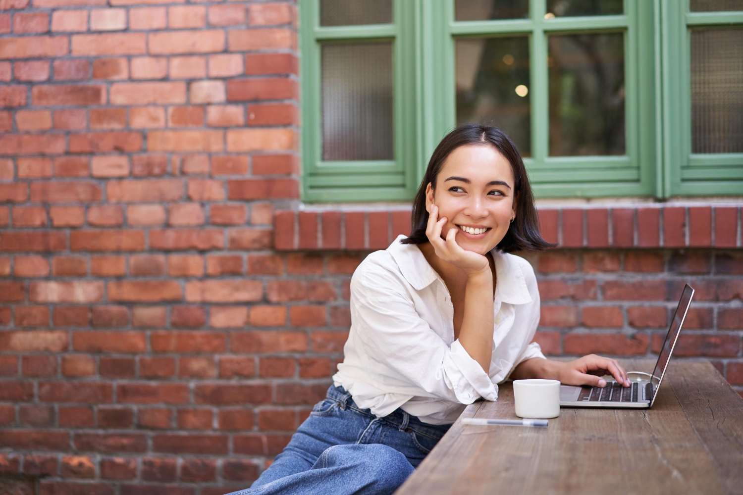Smiling woman sitting at an outdoor table with a laptop and coffee, resting her chin on her hand in front of a brick wall and green window