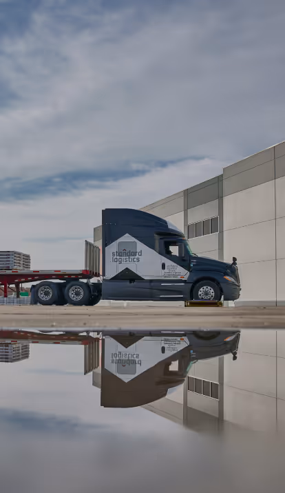 Standard Logistics vehicles near a warehouse, reflected on water.
