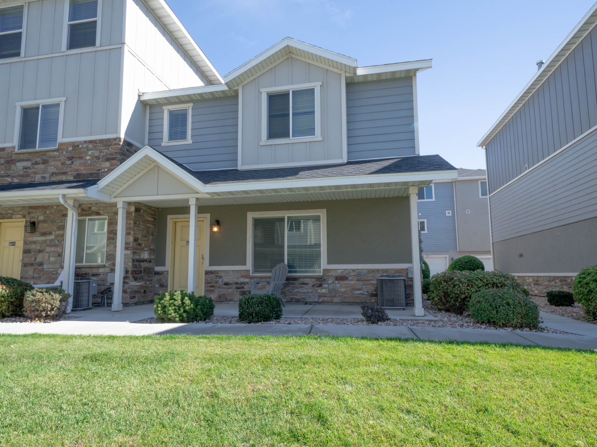 Two-story townhouse with a covered porch, yellow door, bushes, and green lawn under a clear sky.