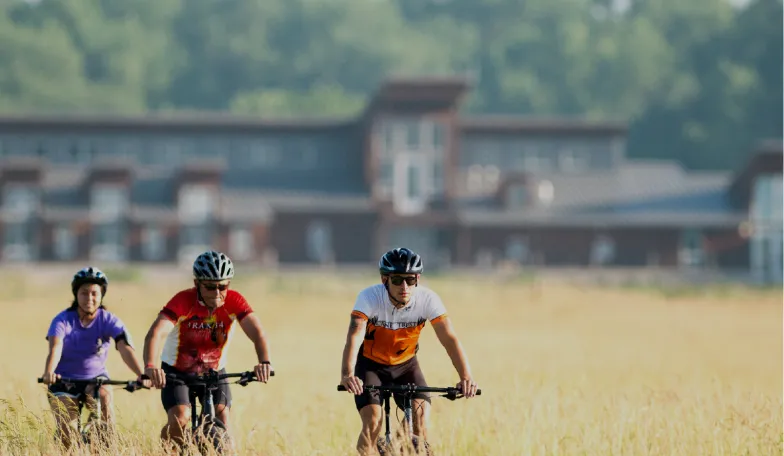 Three cyclists in helmets ride through a grassy field, with a large building in the background. The scene conveys a sense of leisure and adventure.