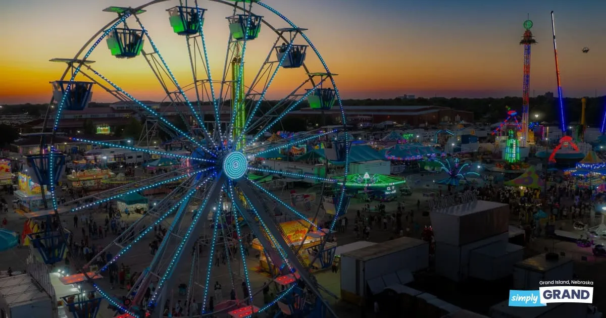 Ferris wheel lit with vibrant lights at sunset, overlooking a bustling fairground with colorful rides and food stalls. The atmosphere is lively.