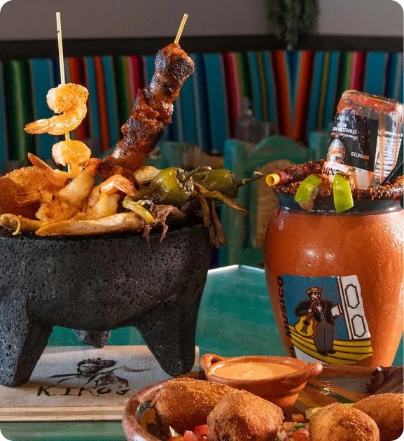 A colorful Mexican dish in a stone bowl with skewered shrimp and grilled vegetables next to a vibrant michelada with an upside-down beer bottle.