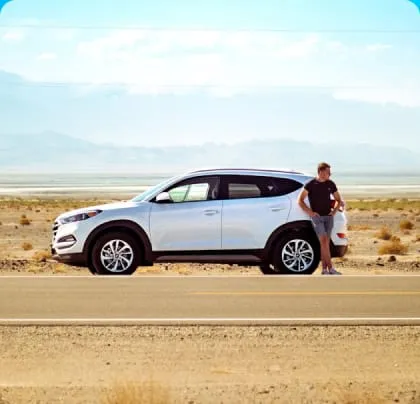 A man leans against a white SUV parked on a deserted desert road under a bright blue sky. The scene conveys a sense of solitude and exploration.