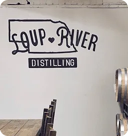 Wall with 'Loup River Distilling' logo depicting Nebraska's outline and heart. Wooden chairs and barrel create a rustic atmosphere.