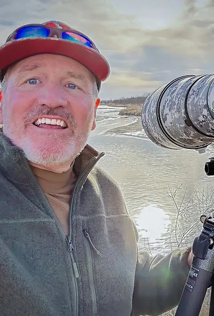 Man smiling beside a large camouflaged camera against a snowy landscape, with a river reflecting sunlight. The scene conveys a sense of adventure and nature appreciation.