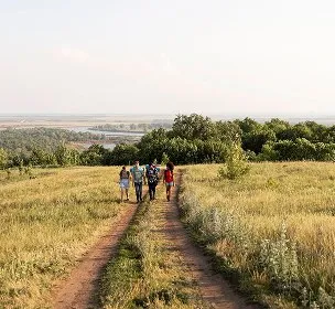 A group of people walks along a dirt path through a sunlit meadow, with lush greenery and a distant river, conveying a sense of adventure and tranquility.