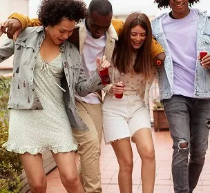 A group of four friends joyfully walks on a rooftop, laughing and holding red cups. They wear casual clothes, embodying a carefree, fun atmosphere.
