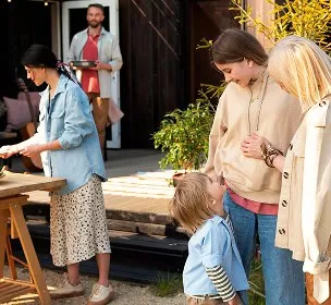 A cheerful family gathers outdoors. A woman and child smile at a pregnant woman, while another woman sets the table, and a man carries food in the background.