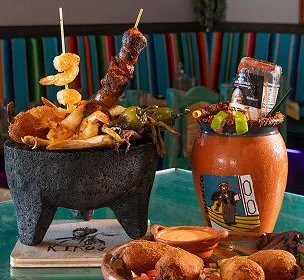 A vibrant platter with skewered meat, shrimp, and peppers in a rustic bowl. A colorful cocktail with an upturned bottle sits beside it. Festive atmosphere.