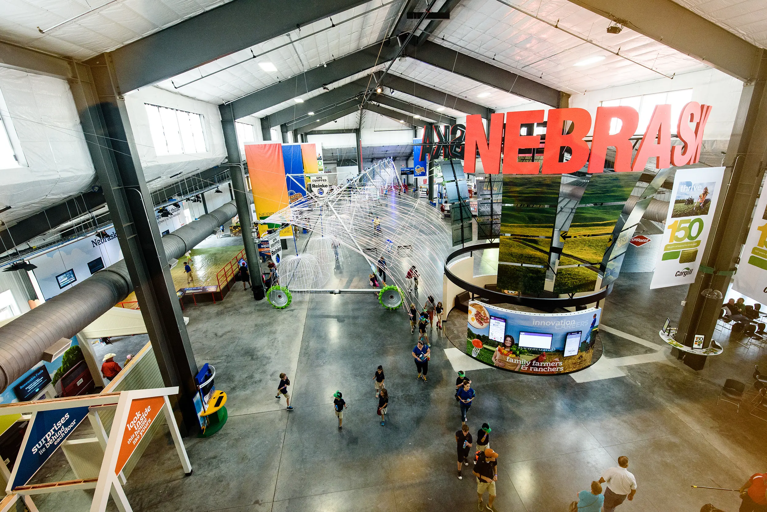Spacious indoor exhibit hall with high ceilings and a large "NEBRASKA" sign. Visitors explore interactive displays and colorful installations. Bright and modern atmosphere.