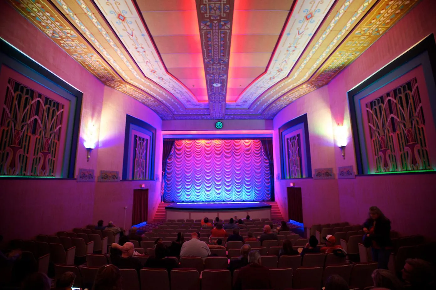 Art deco theater interior with ornate ceiling and colorful lighting, featuring an empty stage with a red curtain. Audience seated, calm atmosphere.
