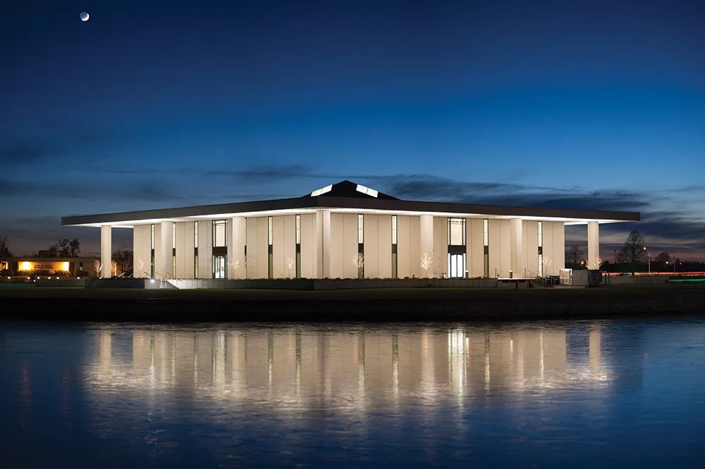 Modern building with tall columns illuminated at night, reflecting on a calm body of water. A crescent moon shines above in a clear, deep blue sky.