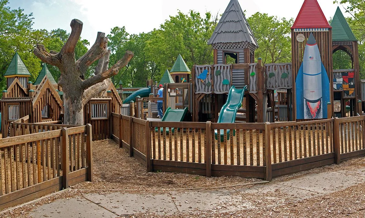 Wooden playground with slide towers, turrets, and a rocket structure, enclosed by a wooden fence. A leafless tree adds a whimsical touch.
