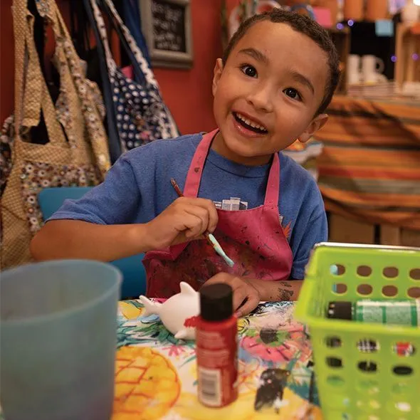 Young child smiling while painting at a colorful table covered with art supplies. The scene is lively, with a warm, creative atmosphere.