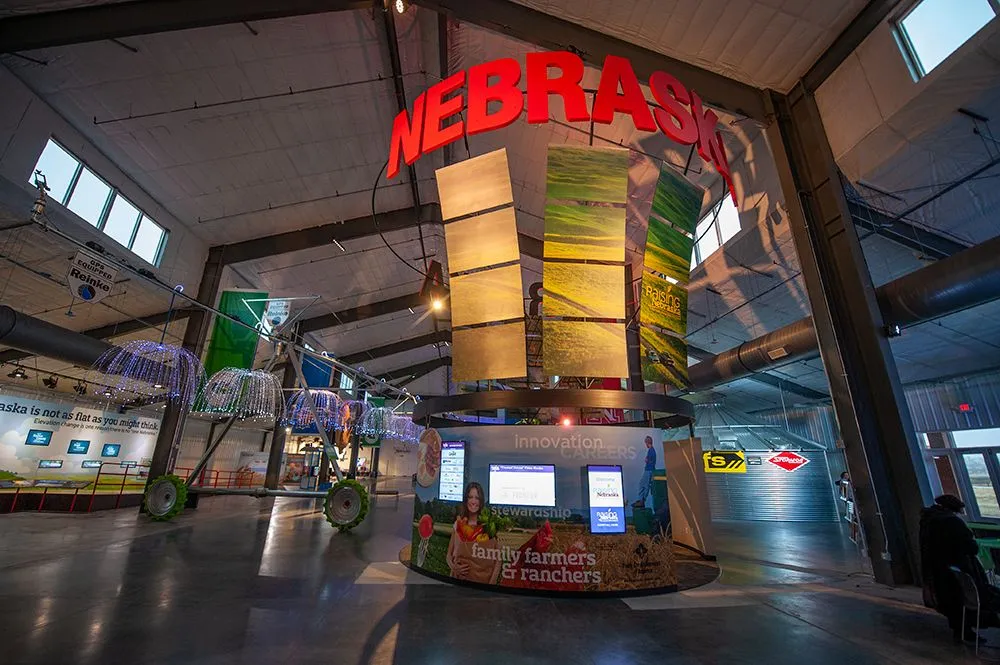 Indoor exhibition with a large "Nebraska" sign above a central kiosk displaying screens. The setup includes bright lights and farming-themed displays.
