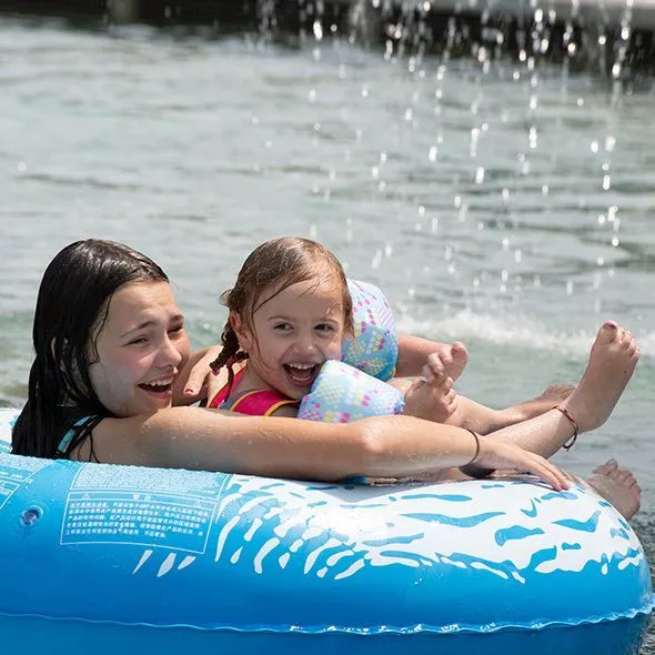 Two smiling girls enjoy a sunny day, floating in a blue inner tube on a sparkling lake, with water splashing playfully around them.