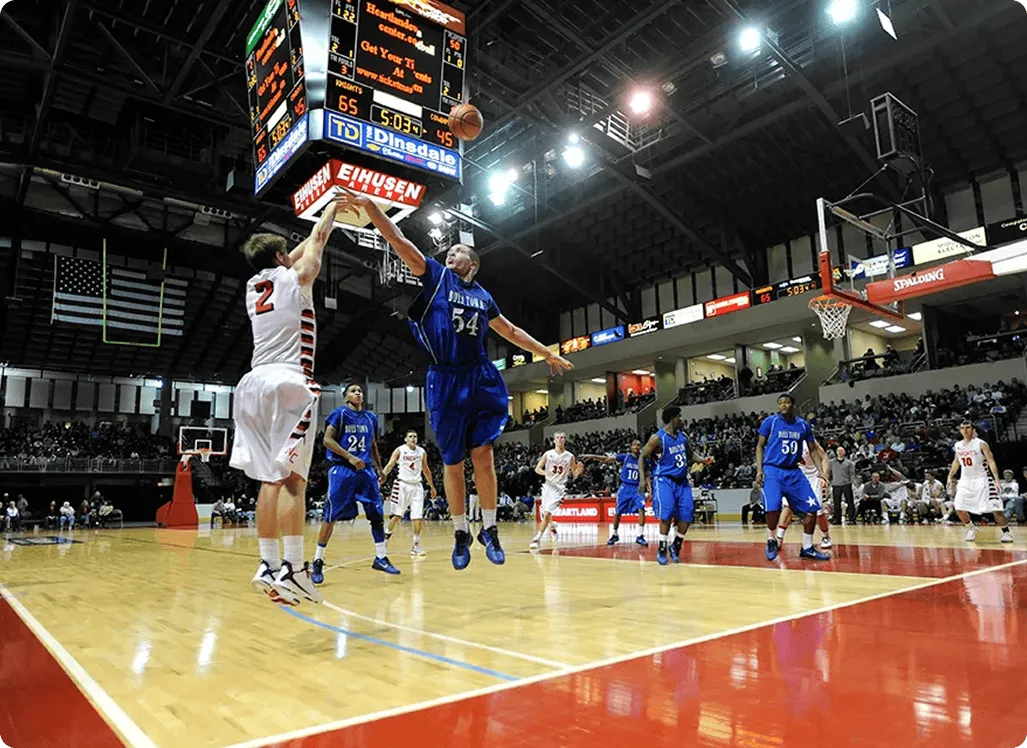A basketball player in a white jersey shoots a jump shot as a defender in a blue jersey attempts to block. The scene is energetic in a crowded indoor arena.