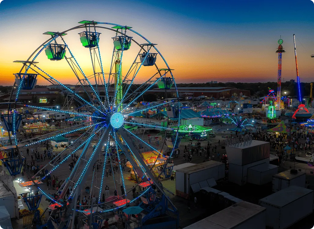 A vibrant carnival scene at sunset, featuring a large, brightly-lit Ferris wheel and colorful booths. The sky is a gradient of orange to deep blue.