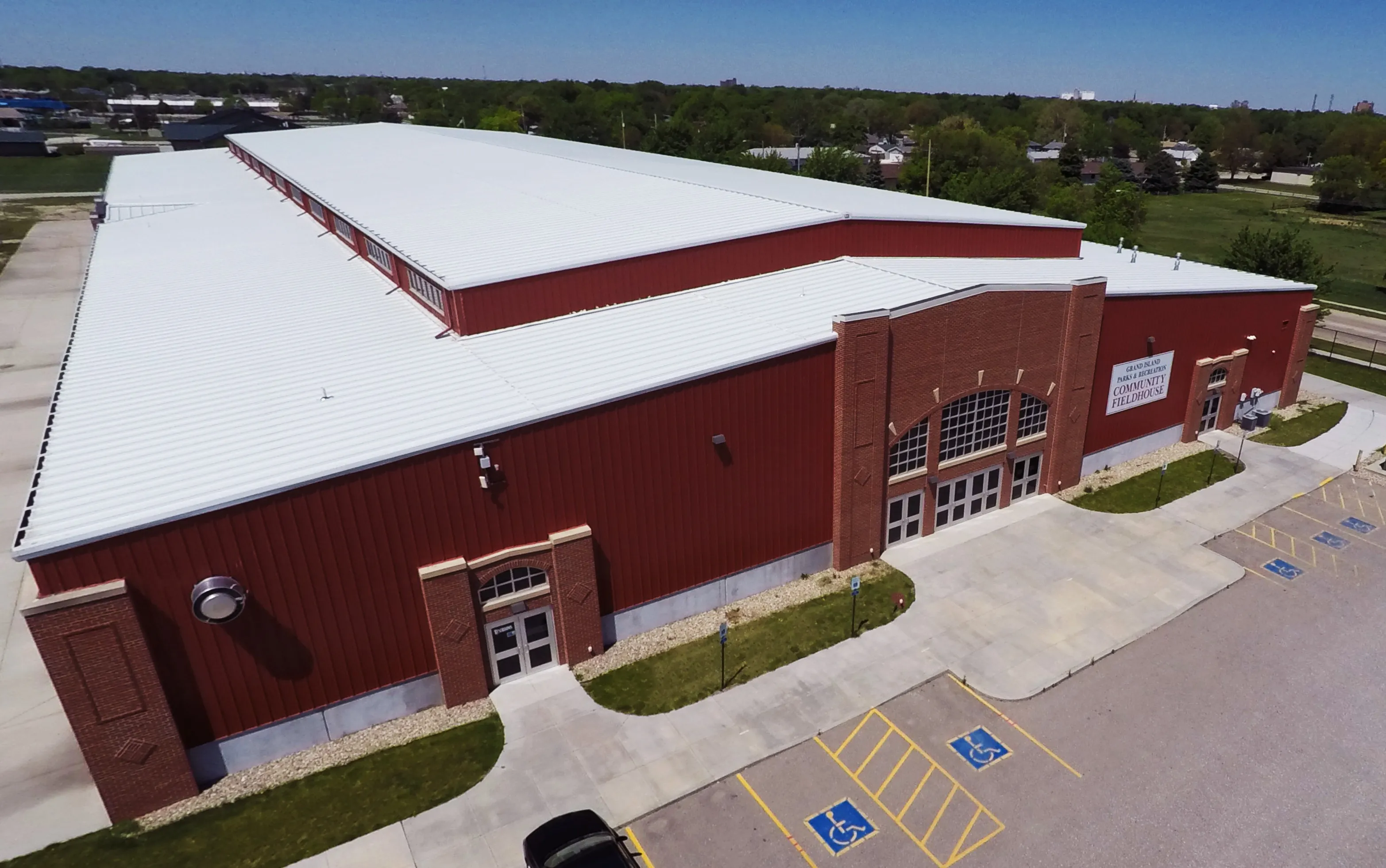 Aerial view of a large red and white warehouse-style building with a metal roof. The surrounding area features parking spaces and green grass.