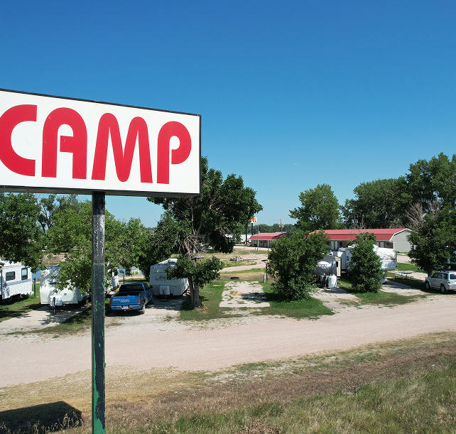 A campsite with a "CAMP" sign in the foreground, surrounded by RVs and trees under a clear blue sky, conveying a peaceful, outdoor vibe.
