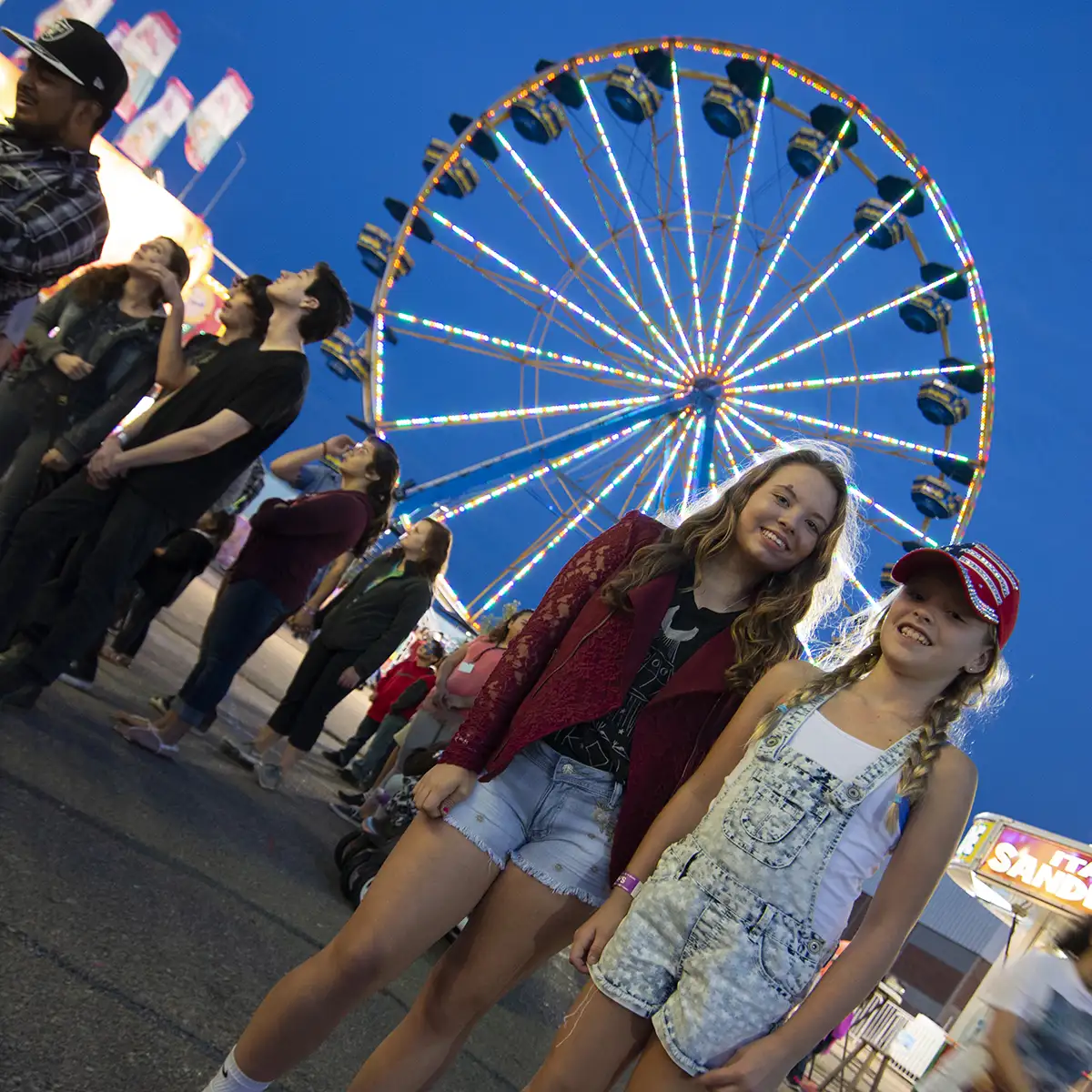 Two young girls stand in front of the Ferris Wheel at the state fair.