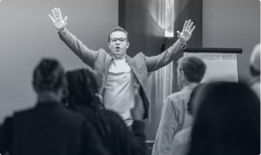 Man in a blazer speaking energetically with raised arms to an audience in a conference room.